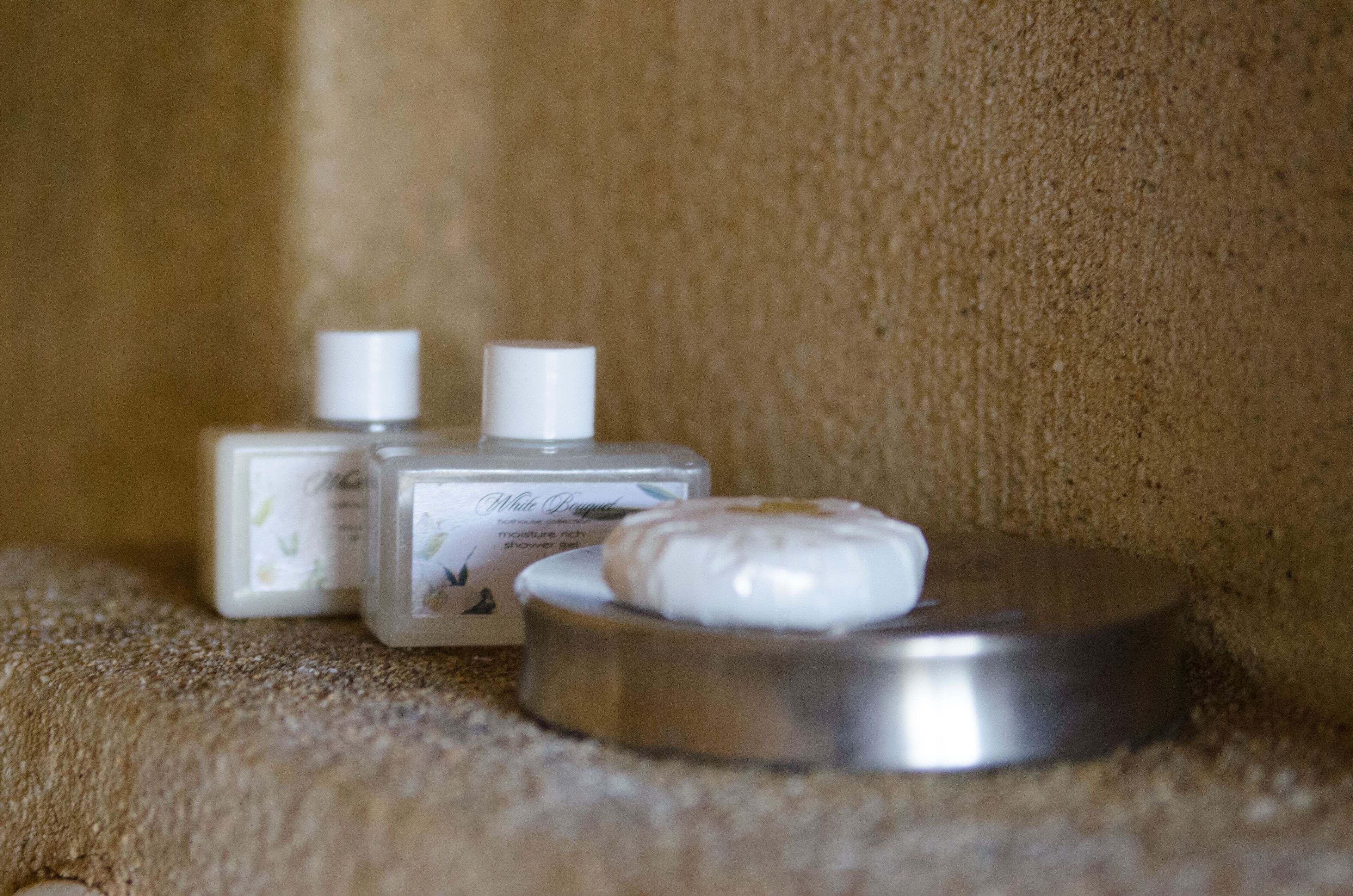 A bathroom with a beige textured wall, a metal tray with a bar of soap, and two bottles of lotion.