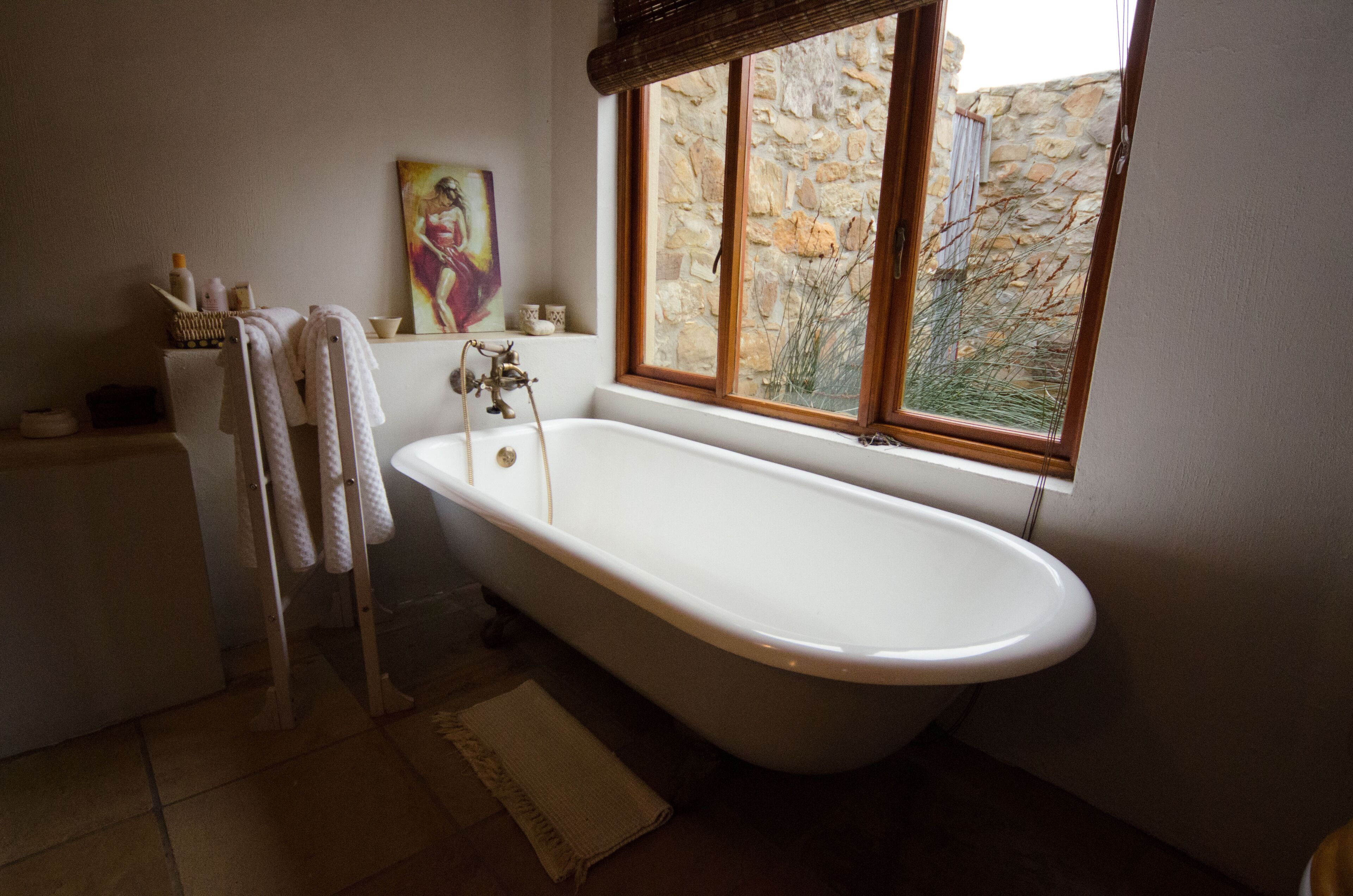 A bathroom with a freestanding bathtub, a window with a stone wall view, and a painting on the wall.