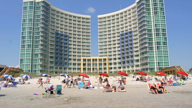 Un gran hotel con varios pisos y balcones, una playa de arena frente a él y varias sombrillas de playa.