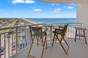 A balcony with a view of the beach, two wooden chairs, a small table, and a railing.