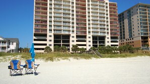 A multi-story hotel with balconies, located on a sandy beach with beach chairs and umbrellas.