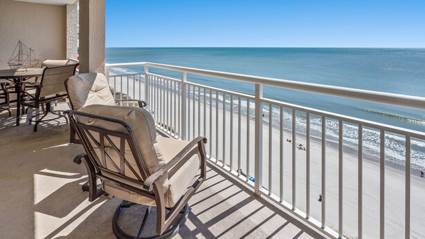 A balcony with a view of the ocean, a table with chairs, and a decorative wall piece.