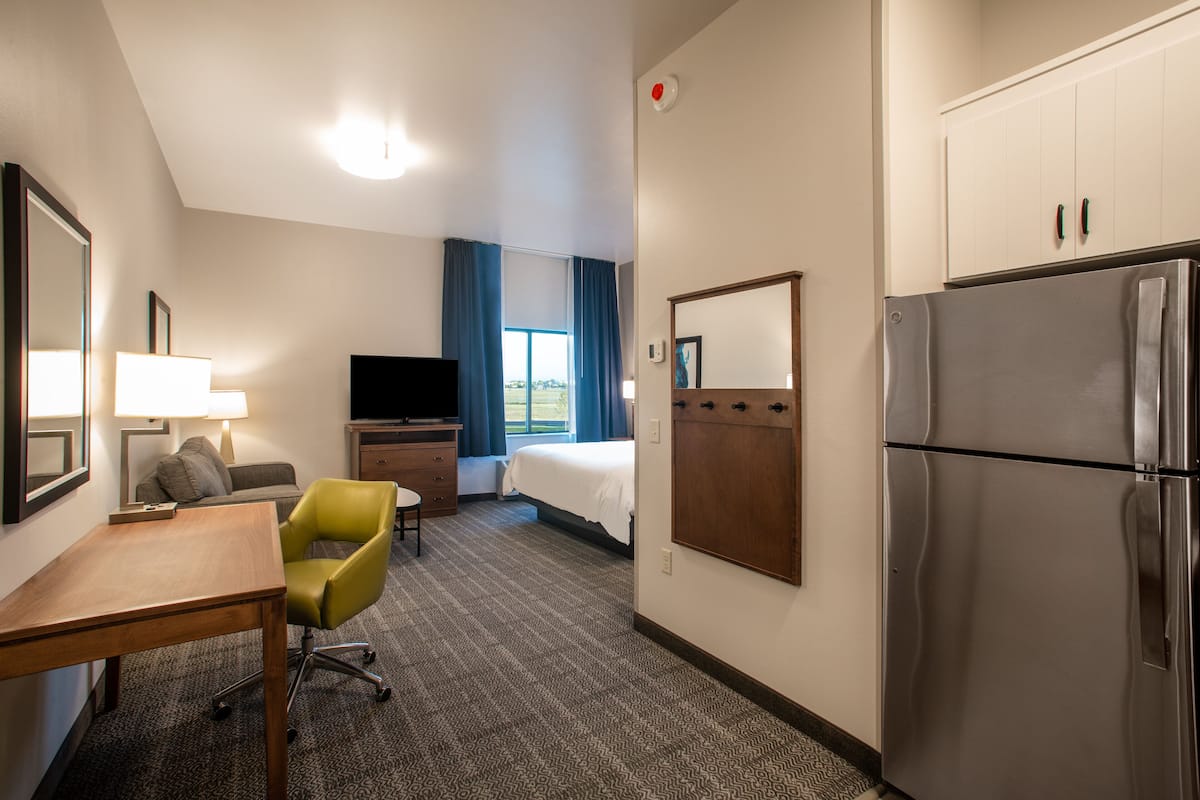 Habitación de hotel con cama, escritorio, silla, televisión, refrigerador y ventana con cortinas.