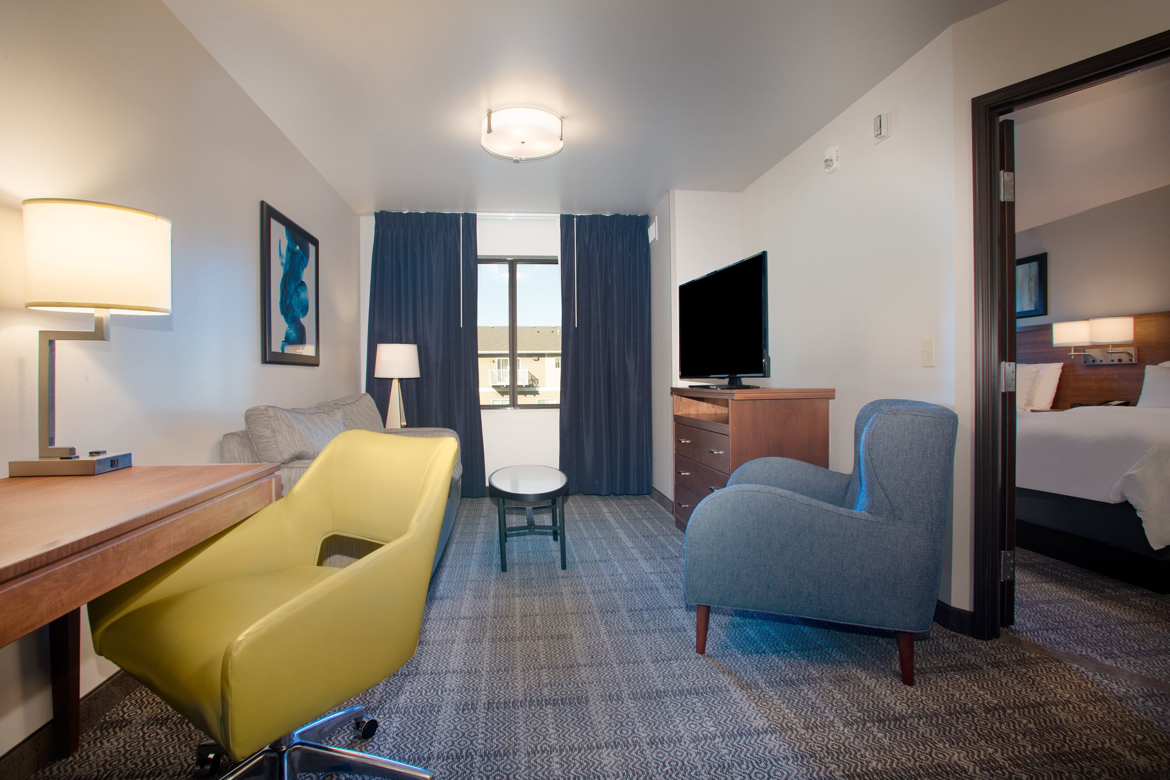 A hotel room with a desk, a yellow chair, a gray armchair, a television, a bed, and a window with curtains.