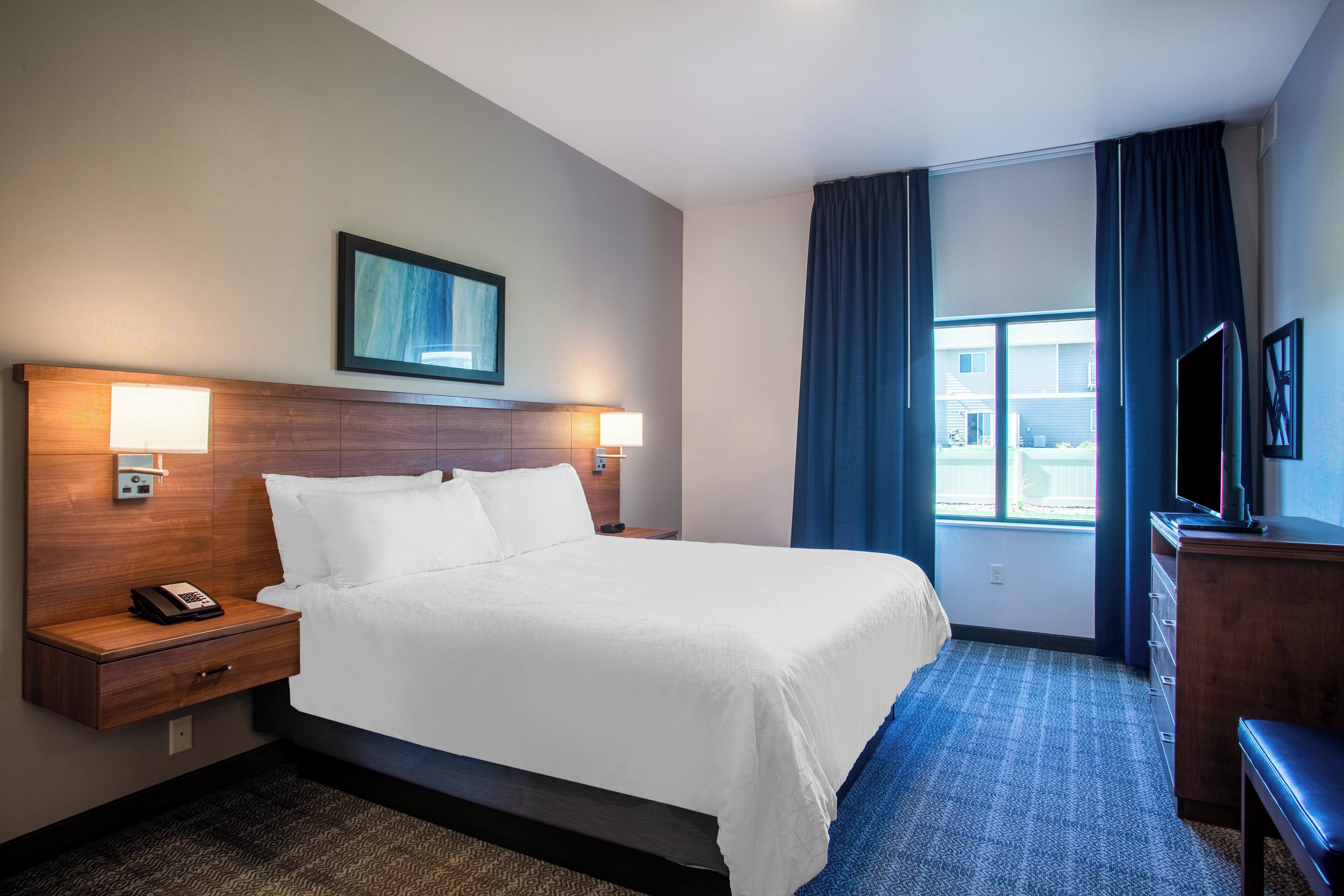 Habitación de hotel con cama, lámparas de noche, teléfono, televisión y ventana con cortinas.