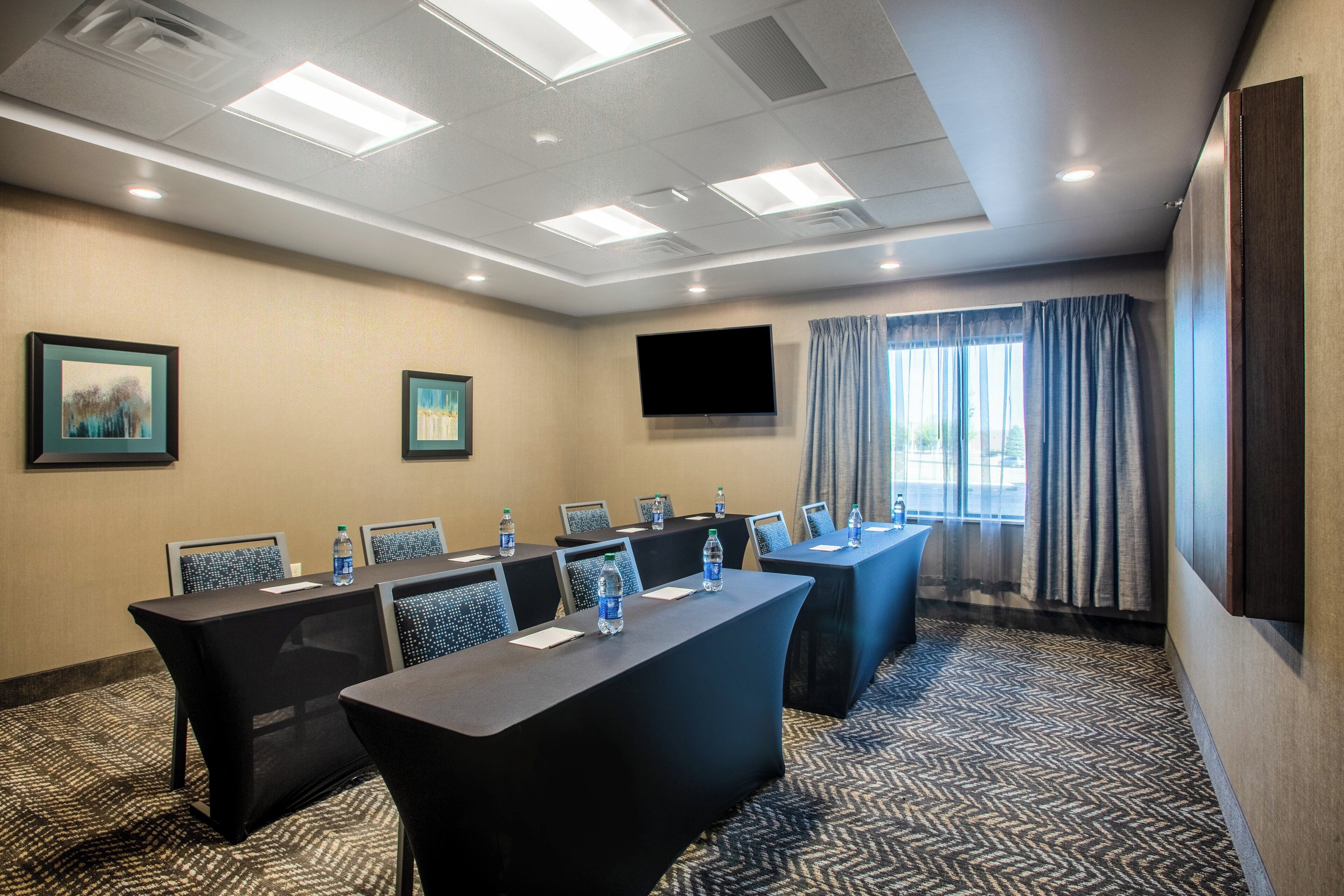 A conference room with a long table, chairs, water bottles, a flat-screen TV, and a window with curtains.