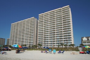 Un hôtel situé en bord de mer, avec plusieurs étages, des balcons et des équipements de plage, notamment des parasols et des chaises longues.