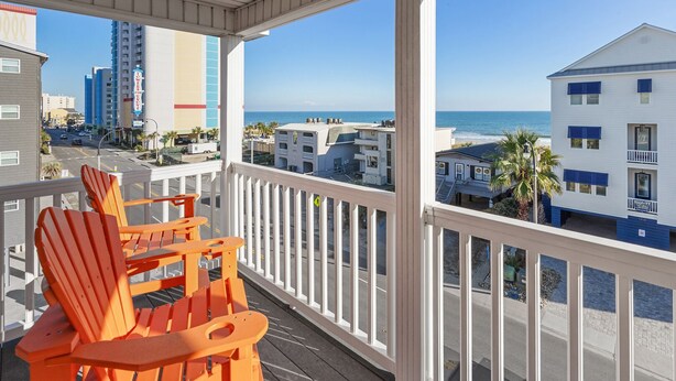 A balcony with orange Adirondack chairs, a white railing, and a view of the beach and ocean.