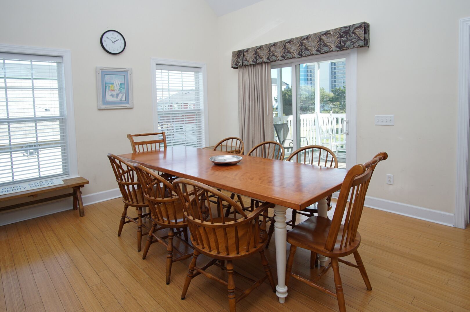 A dining room with a wooden table and six chairs, large windows with blinds, a clock on the wall, and a bench outside the window.