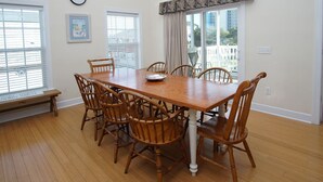 Comedor con una mesa de madera y seis sillas, ventanas amplias con persianas, un reloj en la pared y un banco fuera de la ventana.