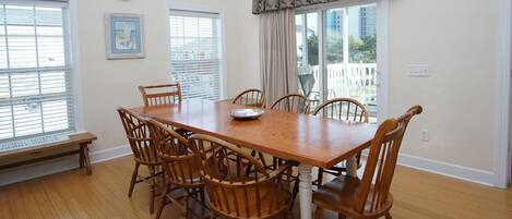 A dining room with a wooden table and six chairs, large windows with blinds, a clock on the wall, and a bench outside the window.