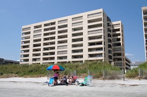 A multi-story building with numerous balconies, a sandy beach area with beach chairs and an umbrella, and a clear blue sky. - Condo with great ocean view, large balcony w/pool & hot tub in popular Cherry Grove Beach (North Myrtle Beach)