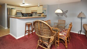 Una cucina compatta con elettrodomestici moderni, una zona pranzo con sedie in vimini e un tavolo in vetro, e un pavimento rivestito di tappeto rosso.