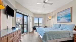 A bedroom with a large bed, a TV, a desk, and a balcony with a view of buildings.