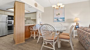 A modern kitchen with built-in appliances, a dining area with wicker chairs and a patterned sofa, and a beach-themed painting on the wall.