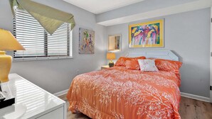 Un dormitorio con cama, escritorio, lámpara, ventana con persianas y un cuadro en la pared.