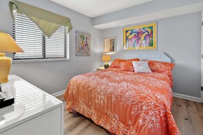 Une chambre à coucher avec un lit, un bureau, une lampe, une fenêtre avec des stores et un tableau au mur.