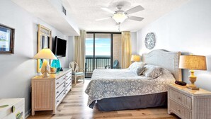 Un dormitorio con cama, mesitas de noche, televisor, ventilador de techo y vistas al exterior.