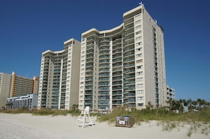 A high-rise hotel with multiple balconies, located near a beach with a lifeguard stand and a beachside sign. - Large oceanfront family-sized condo rental w/lazy river, pool, and fitness center near Ocean Drive (North Myrtle Beach)
