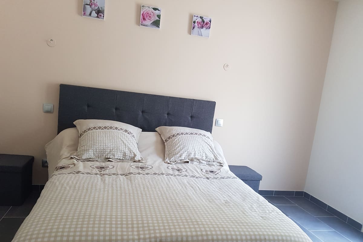 Ein ordentlich bezogenes Bett mit einem karierten Überwurf und zwei Kissen, ein dunkles Kopfstück und eine Wand mit drei kleinen floralen Bildern.
