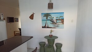 Een klein eetgedeelte met een houten stoel, een groene tafel en krukken. Op de muur hangt een schilderij van een strandgezicht.