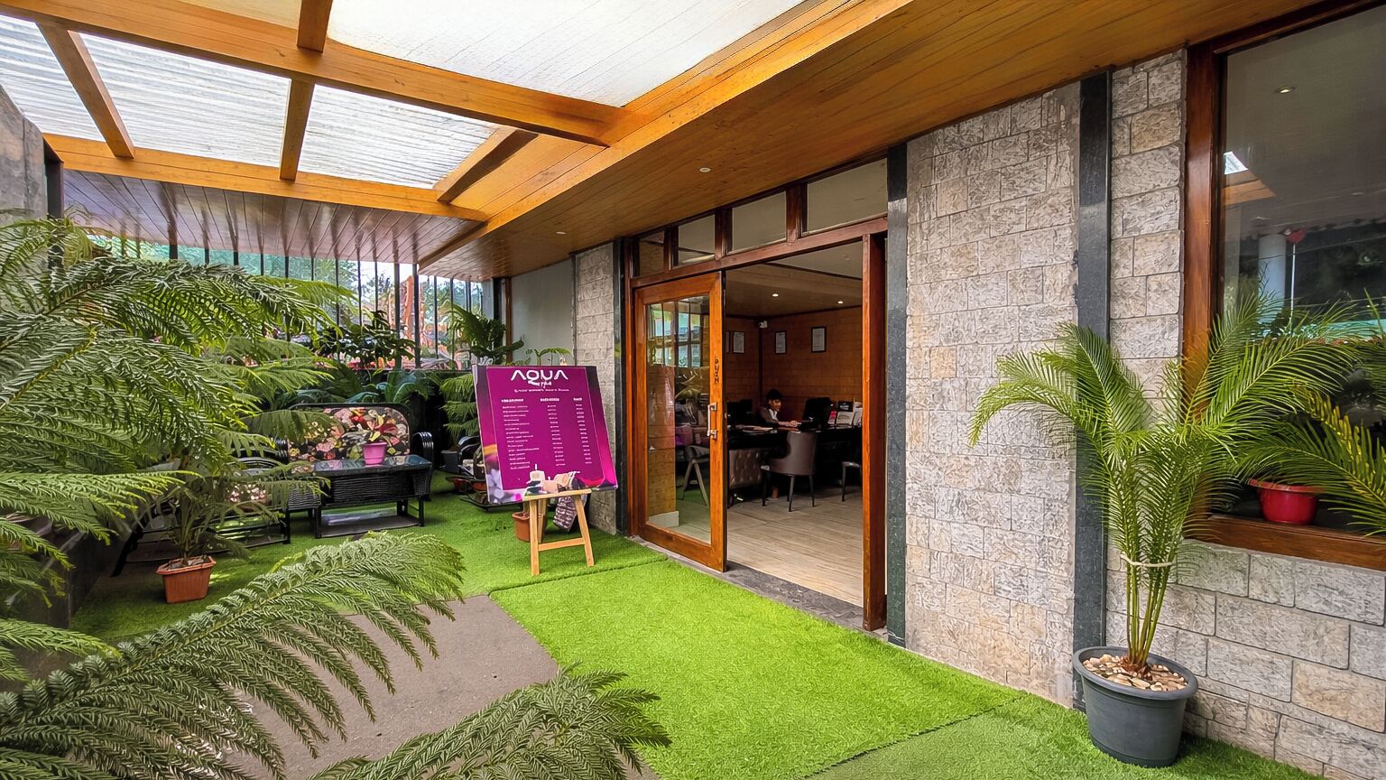 A hotel lobby with a green artificial turf, potted plants, a wooden ceiling, stone walls, and a glass door leading inside.