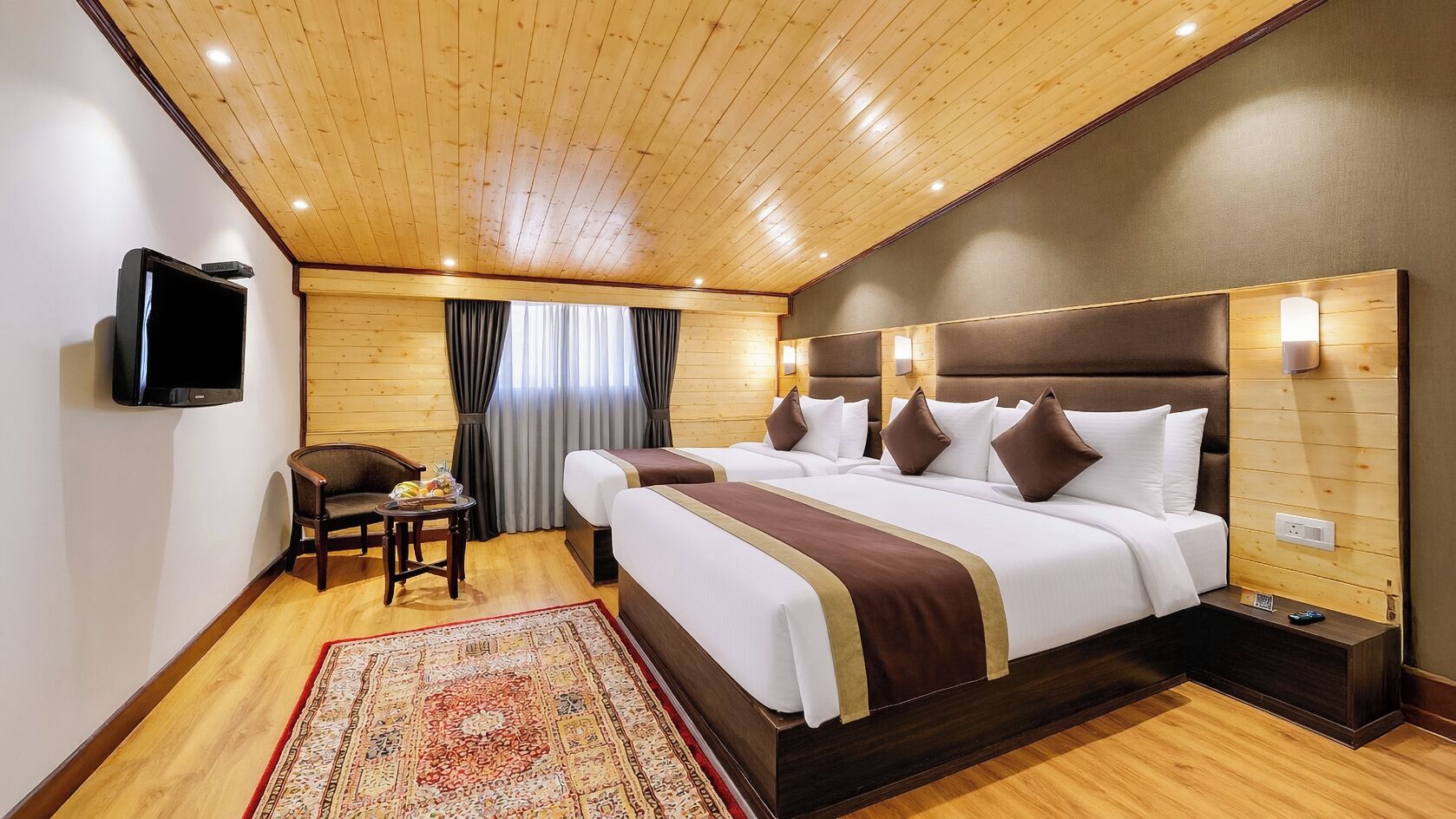 A hotel room with a wooden ceiling, a large bed with multiple pillows, a TV, a small table with a chair, and a decorative rug.