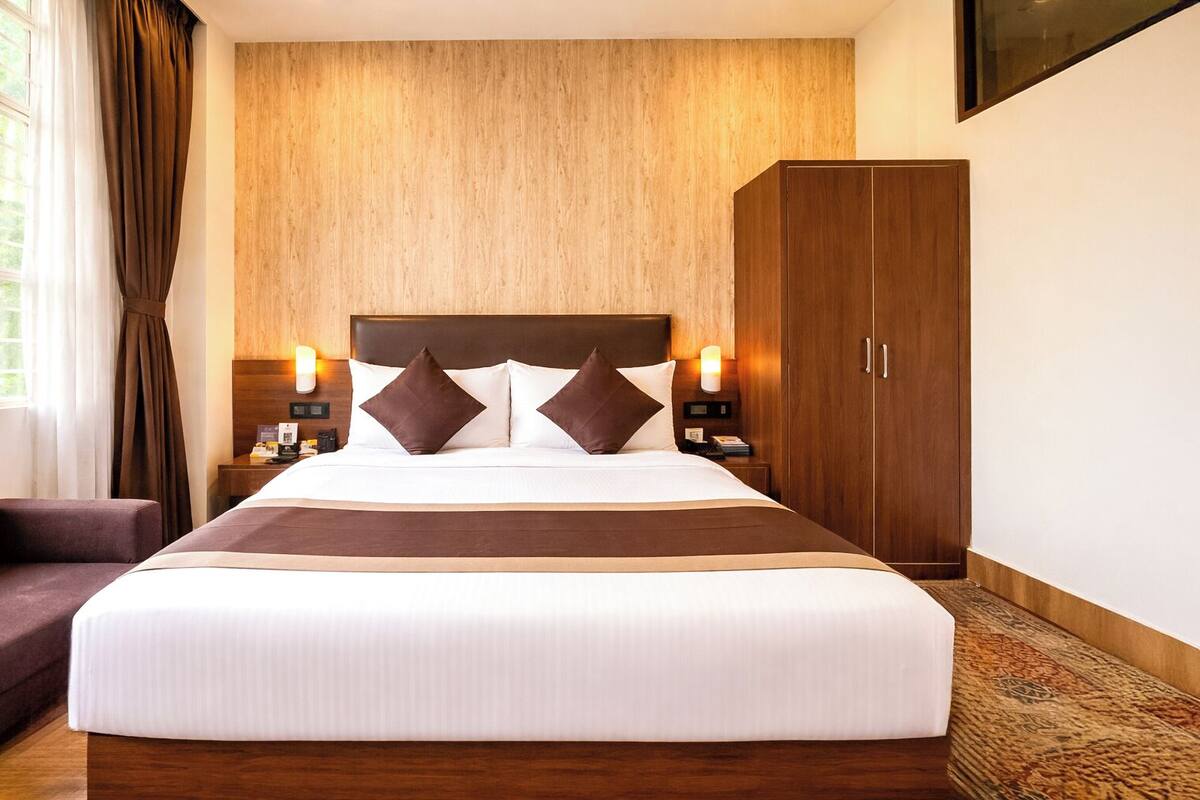 A hotel room with a large bed, a wooden headboard, a nightstand with a lamp, a wardrobe, and a window with curtains.