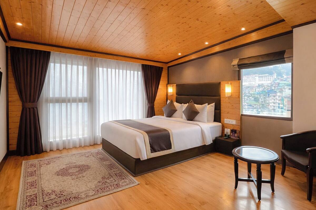 A hotel room with a wooden ceiling, a large bed, a bedside table, a chair, and a window with a city view.