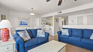 A living room with two blue sofas, a white coffee table, and a red vase. There is a ceiling fan and a framed picture on the wall.