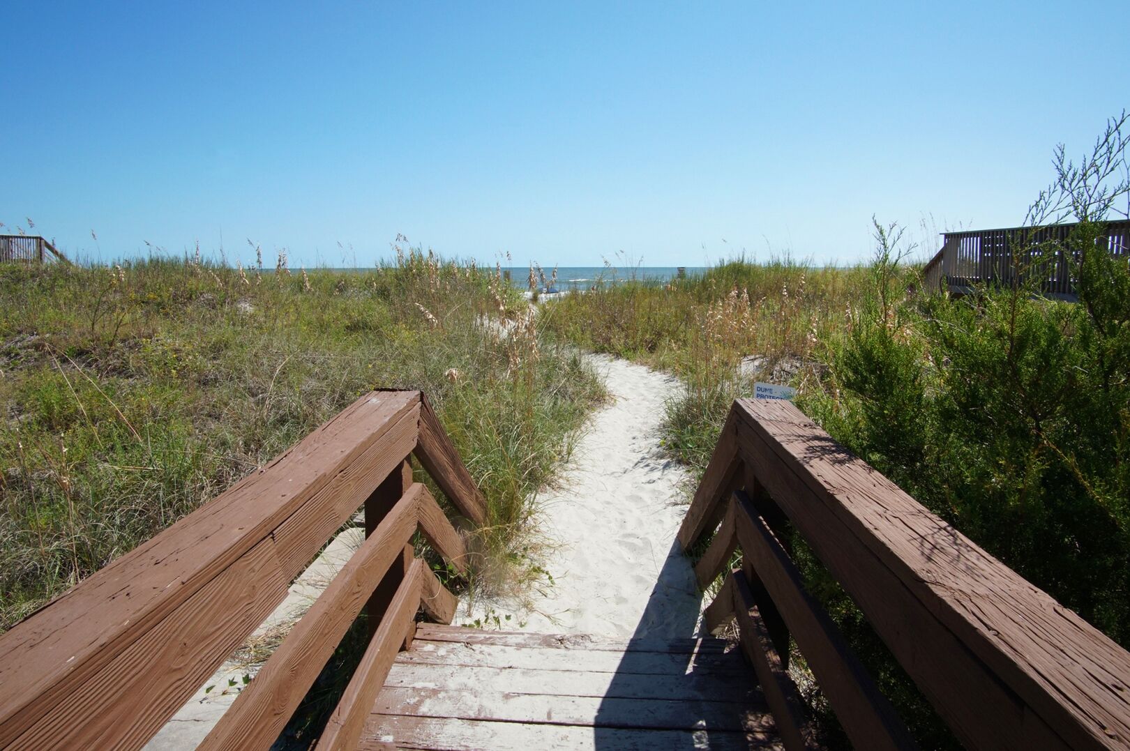 A wooden boardwalk leading to a beach with sand and grassy dunes.