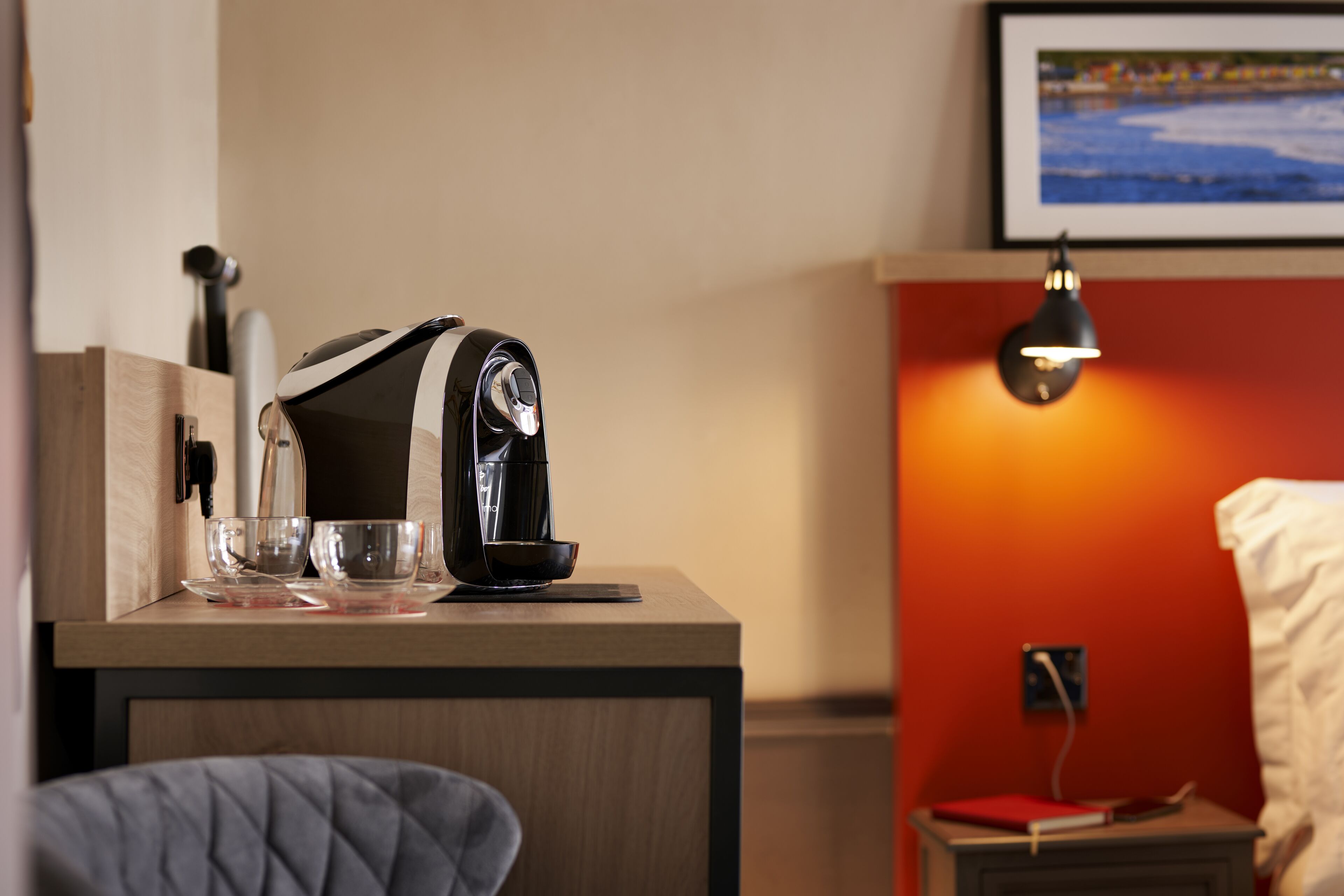 Et hotelværelse med et træskrivebord, en kaffemaskine, en skål og et natbord med lampe.