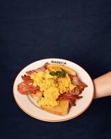 A plate of scrambled eggs, bacon, and toast with a garnish, labeled "BARECA".