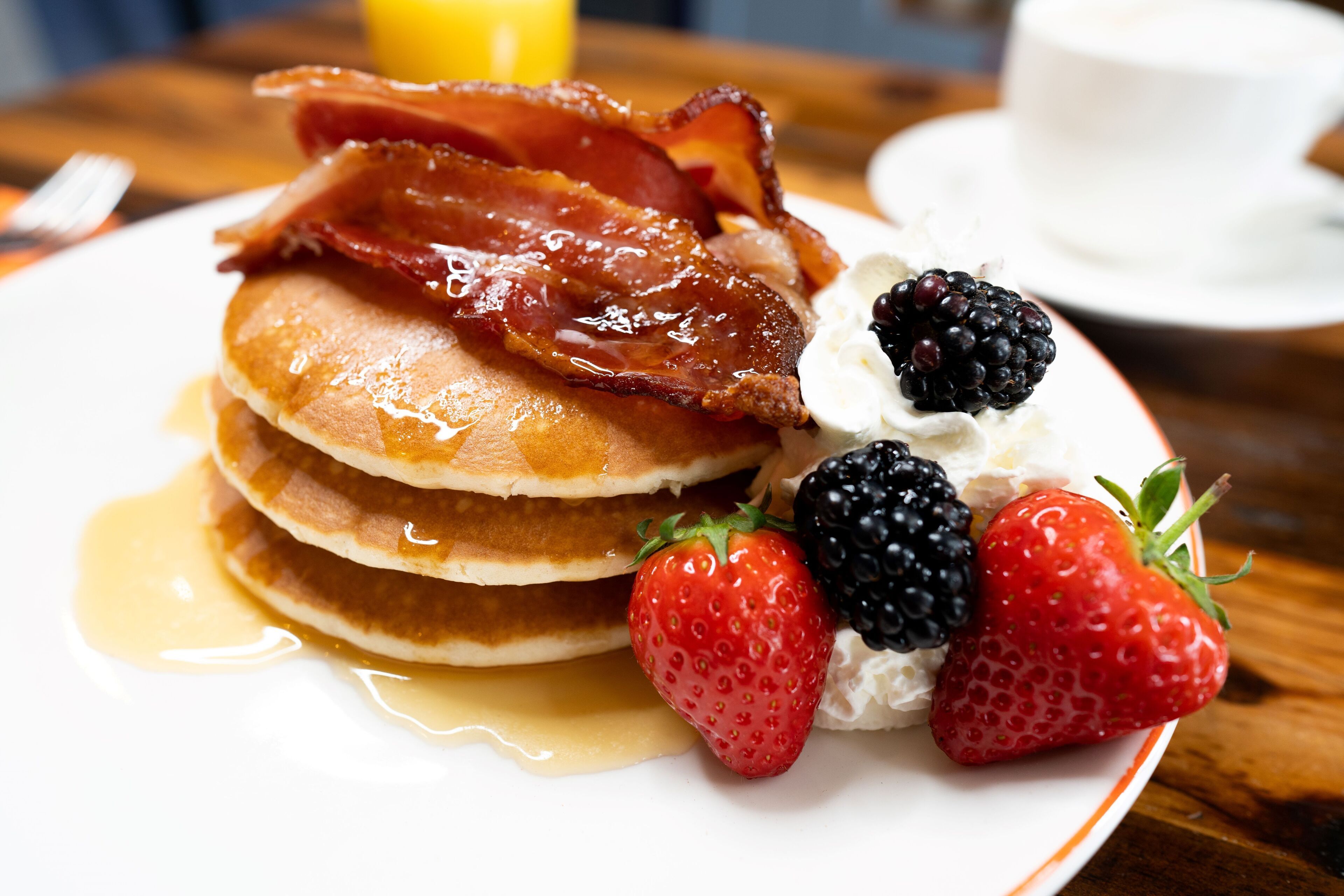 Un plato de tortitas con tocino, fresas, frambuesas, crema batida y jarabe.