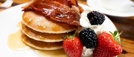Un plato de tortitas con tocino, fresas, frambuesas, crema batida y jarabe.
