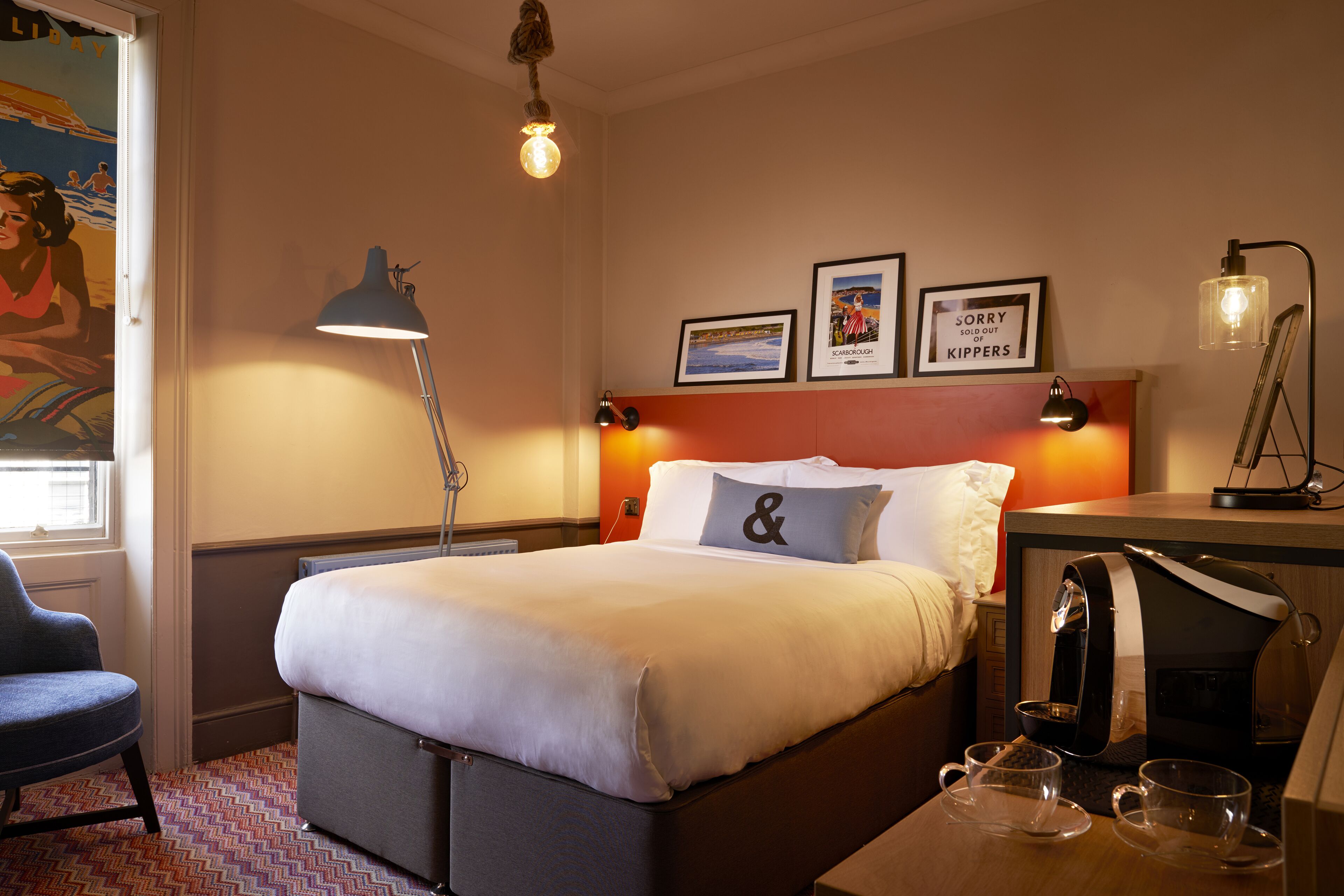 A hotel room with a bed, a desk with a coffee maker, a chair, and a lamp.