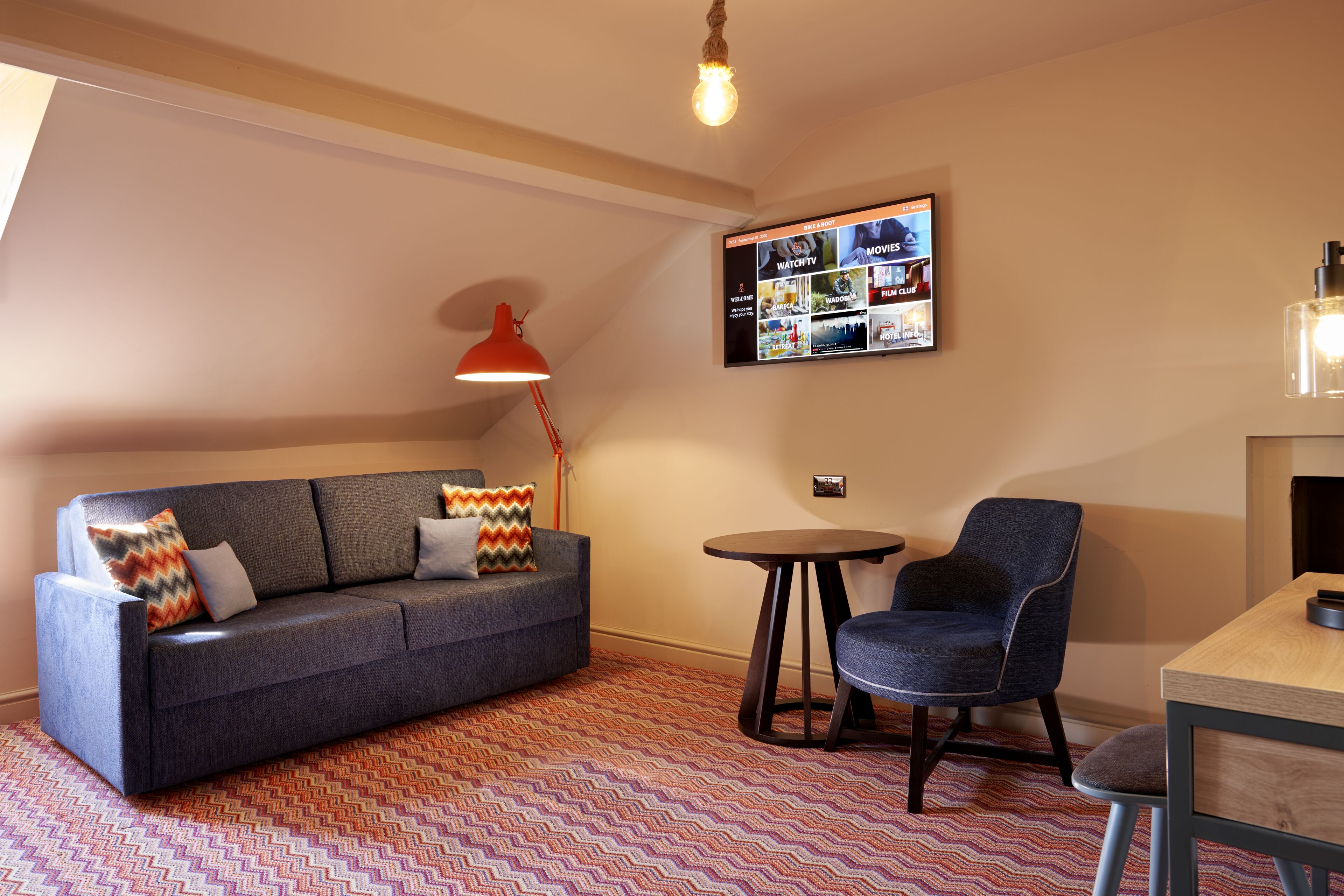 A modern living room with a sofa, a chair, a small table, and a TV mounted on the wall displaying various channels.