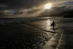 Escena de playa con una persona cargando una tabla de surf, olas y un atardecer.
