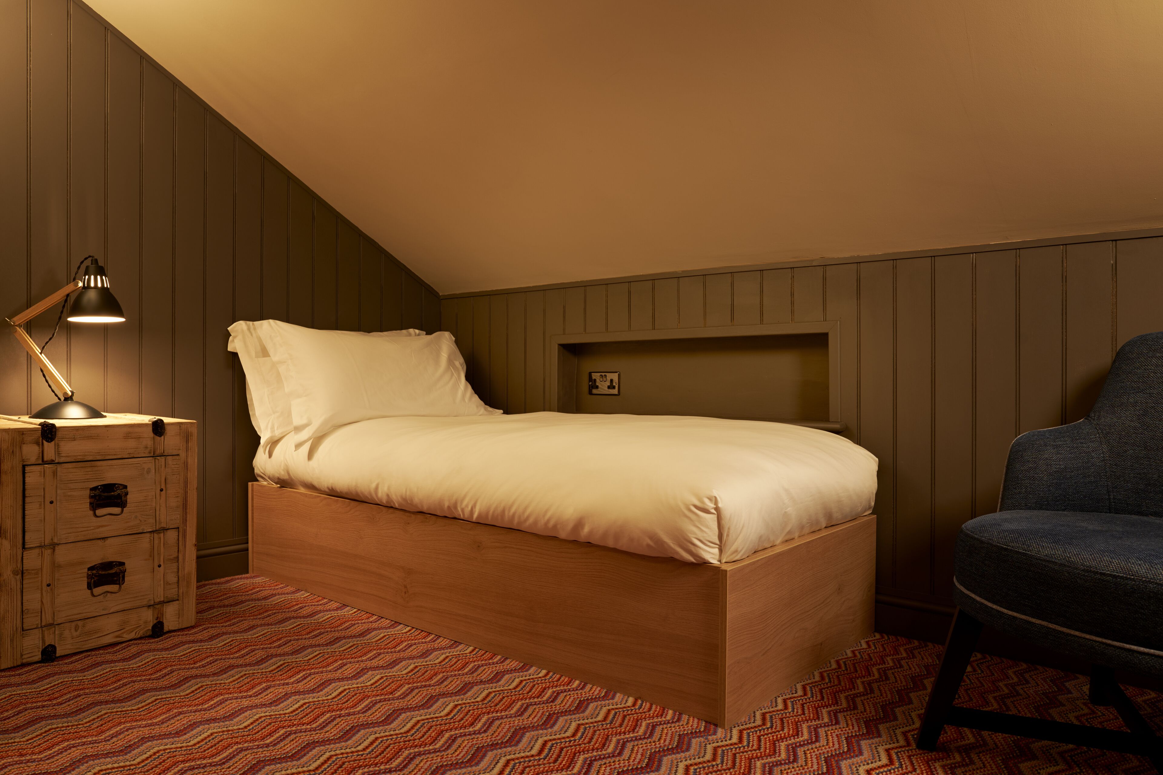 Een eenpersoonsbed met houten frame, een bedlamp, een stoel en een kamer met houten lambrisering en een kamerbreed tapijt met dessin.