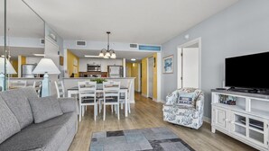 Una sala moderna con un sofá gris, una zona de comedor con sillas blancas, un mueble de televisión con libros y una cocina al fondo.