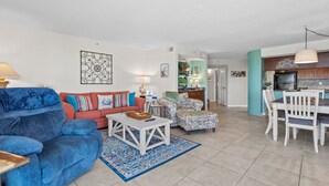 A spacious living room with a blue sofa, a red sofa, and a floral armchair. There is a dining area with white chairs and a table. The kitchen is visible in the background. - Spacious, oceanfront family condo rental w/amazing views, pool, hot tubs in North Myrtle Beach (North Myrtle Beach)