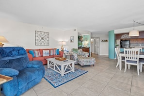 Ein geräumiges Wohnzimmer mit einem blauen Sofa, einem roten Sofa und einem blumigen Sessel. Es gibt eine Essecke mit weißen Stühlen und einem Tisch. Im Hintergrund ist die Küche zu sehen.