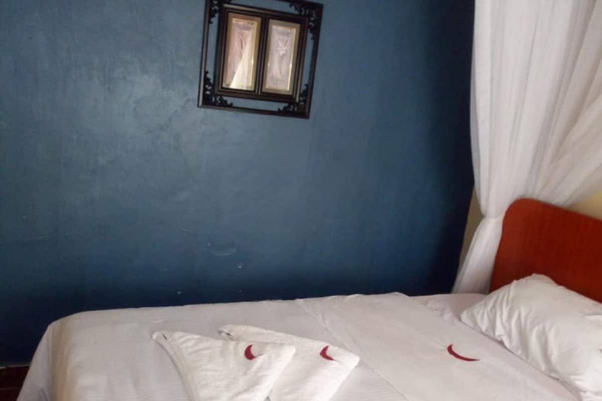 A bed with white linens and towels arranged on it, a blue wall, a framed picture, and a white curtain.