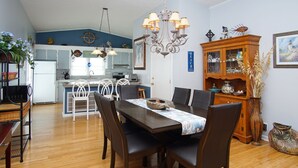 Uma sala de jantar com uma mesa e cadeiras de madeira, um lustre e uma cozinha ao fundo.