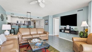 A spacious living room with a beige leather sofa, a glass coffee table, and a colorful rug. A kitchen with white cabinets and a bar area is visible in the background. - Premier resort offering deluxe oceanfront condos in the heart of family-friendly North Myrtle Beach (North Myrtle Beach)