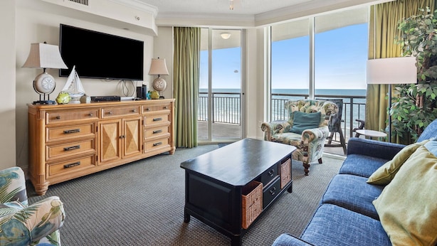 A spacious living room with a large flat-screen TV, wooden entertainment unit, and a balcony with a view of the ocean. - Luxe oceanfront resort with pools, lazy river, hot tubs, spa, fitness center, poolside bar (North Myrtle Beach)