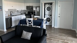 A compact kitchen with white cabinets, a microwave, and a dishwasher. There is a dining area with a table and chairs, and a black couch with a patterned pillow. A washing machine is visible in the background.