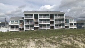 A multi-story beachfront building with balconies, a sandy foreground, and a cloudy sky.