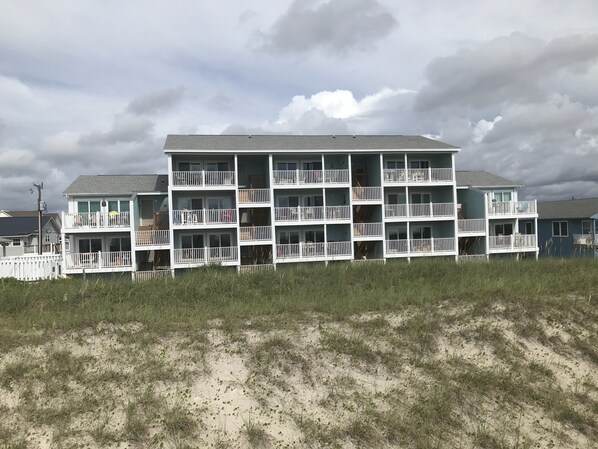 A multi-story beachfront building with balconies, a sandy foreground, and a cloudy sky.