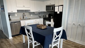 A modern kitchen with white cabinets, a stainless steel dishwasher, a microwave, and a black refrigerator. There is a dining table with a dark tablecloth and white chairs.